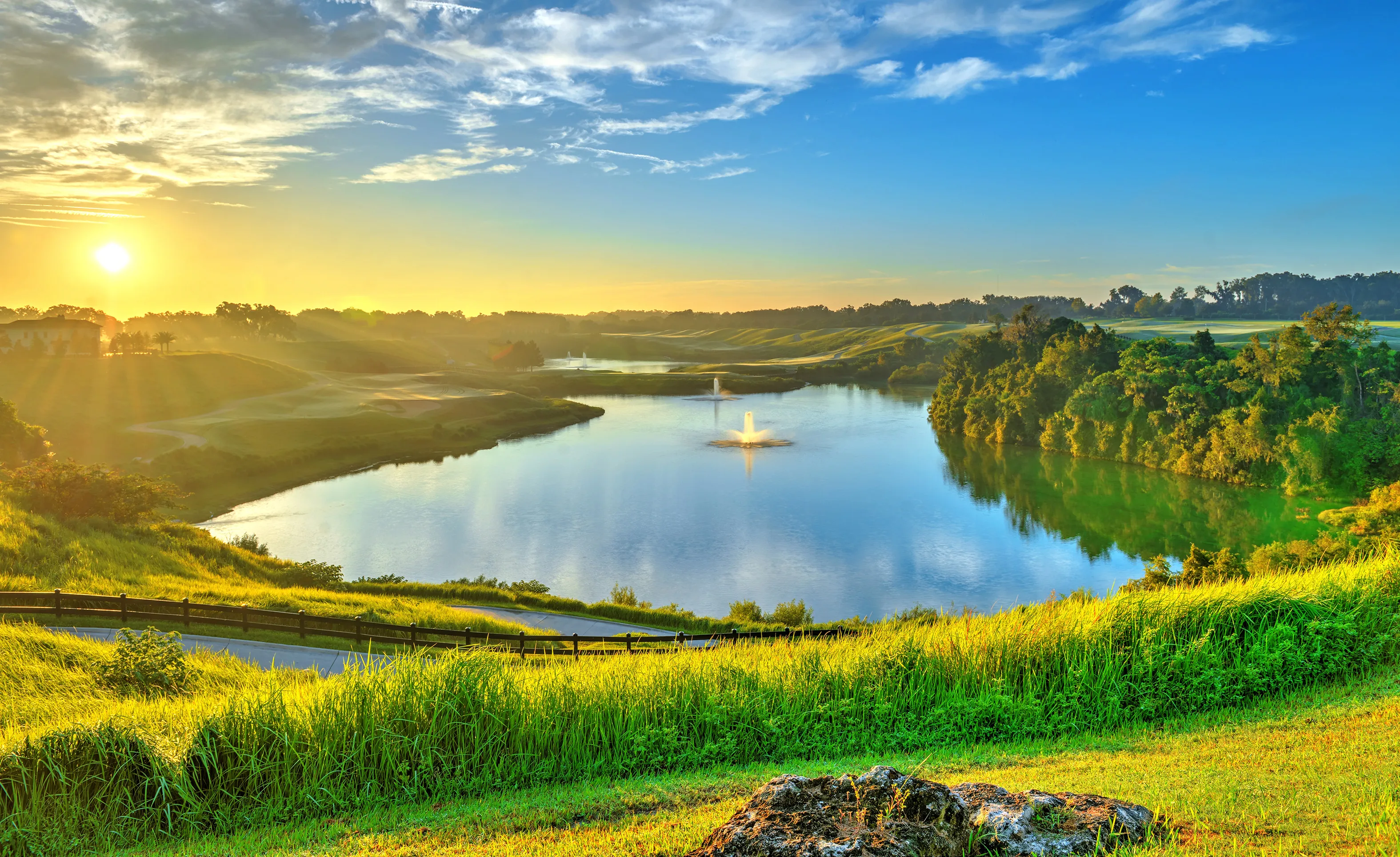Sunrise over a calm lake with water fountains, surrounded by green hills and trees in the IronCrest community.