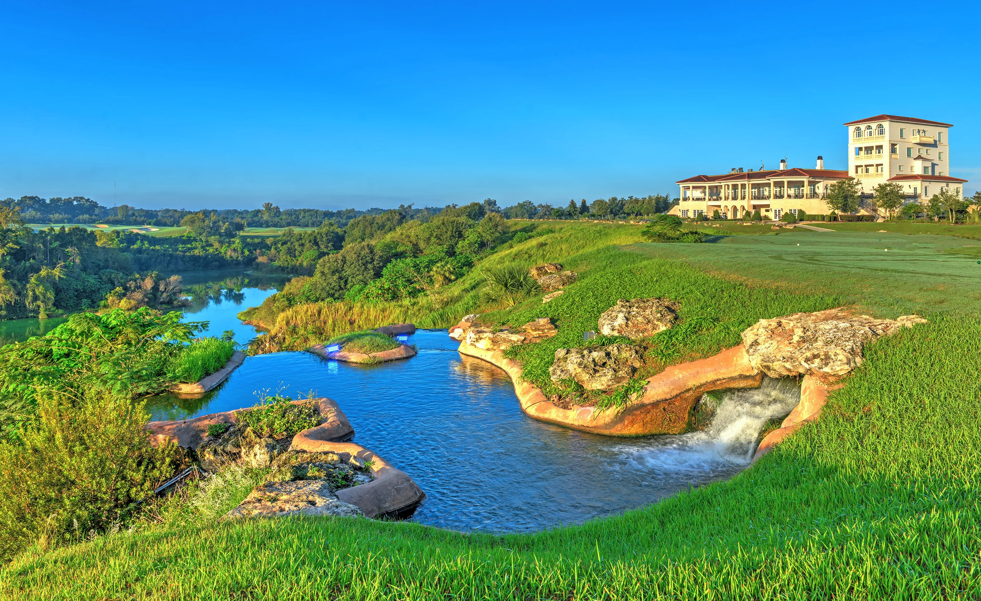 A large multi-story clubhouse overlooks a scenic golf course, pond, and landscaped waterfall in the IronCrest community.