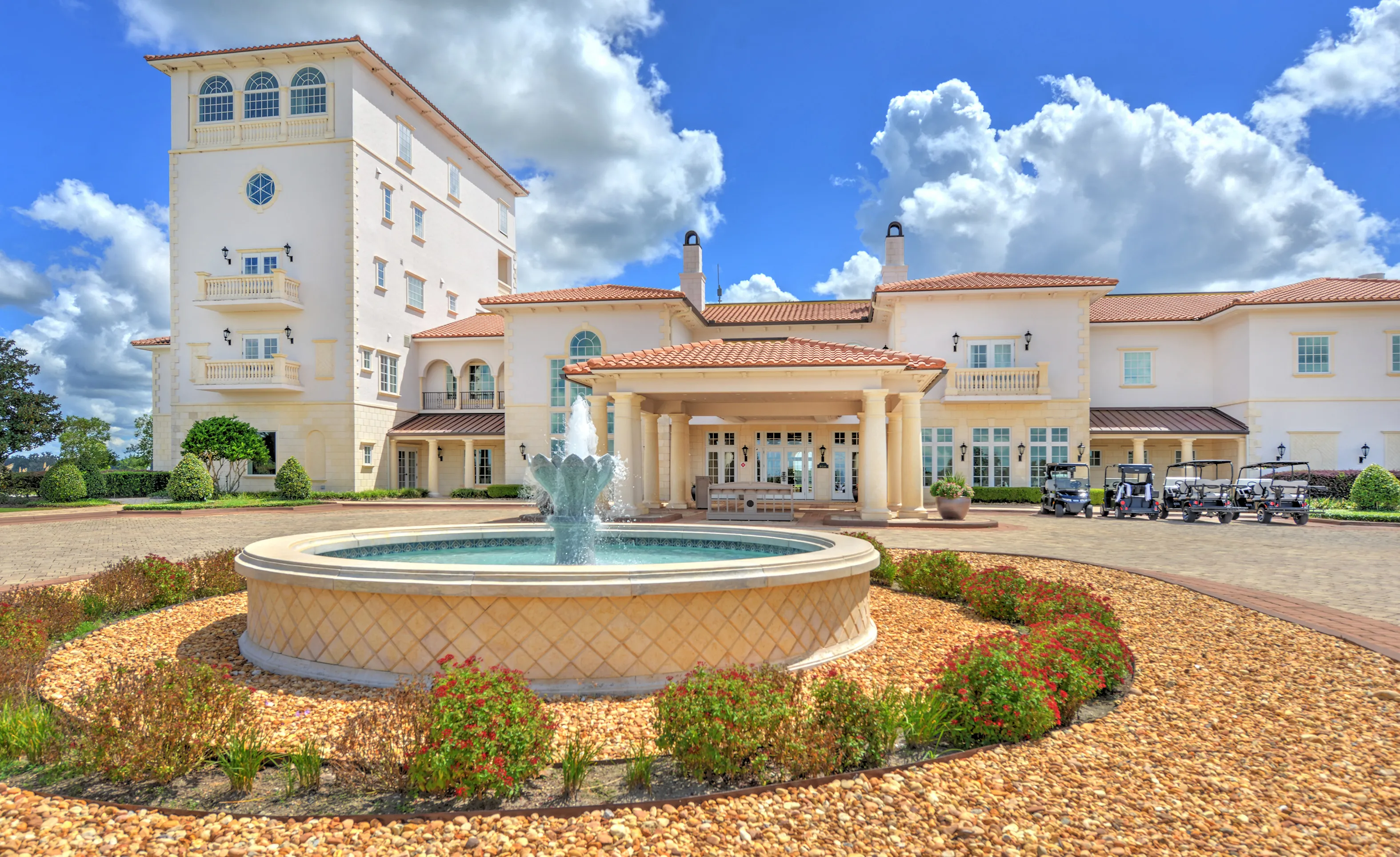 Large Mediterranean-style clubhouse with a multi-story tower, tiled roof, and a central circular fountain in the front.
