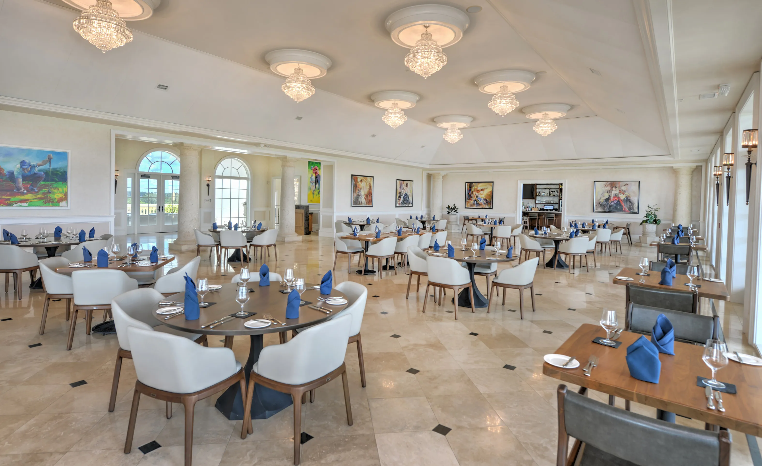 Spacious clubhouse dining room with vaulted ceilings, crystal chandeliers, stone pillars, and multiple tables with blue napkins.