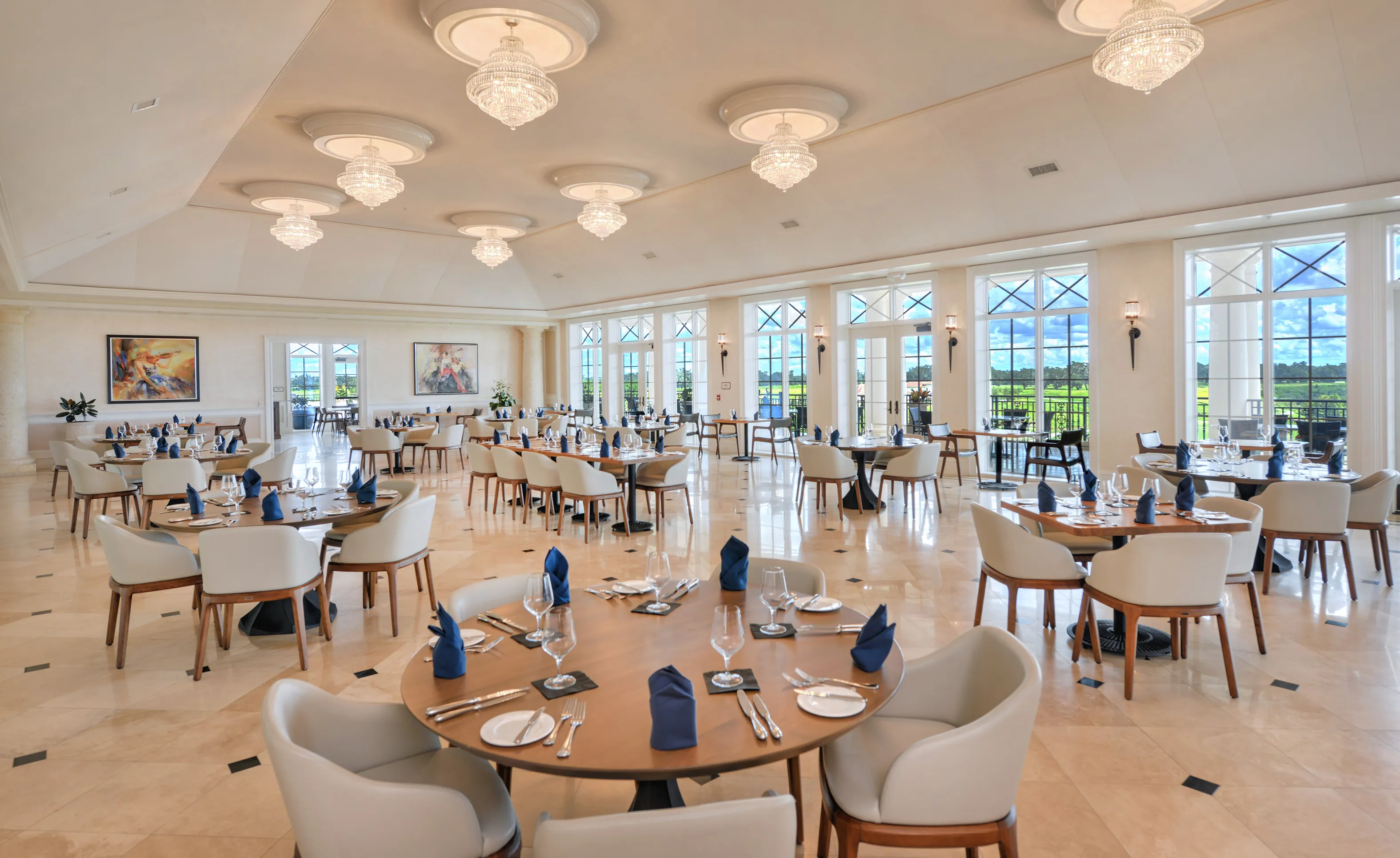 Spacious banquet hall with round tables, light-colored armchairs, crystal chandeliers, and floor-to-ceiling windows.