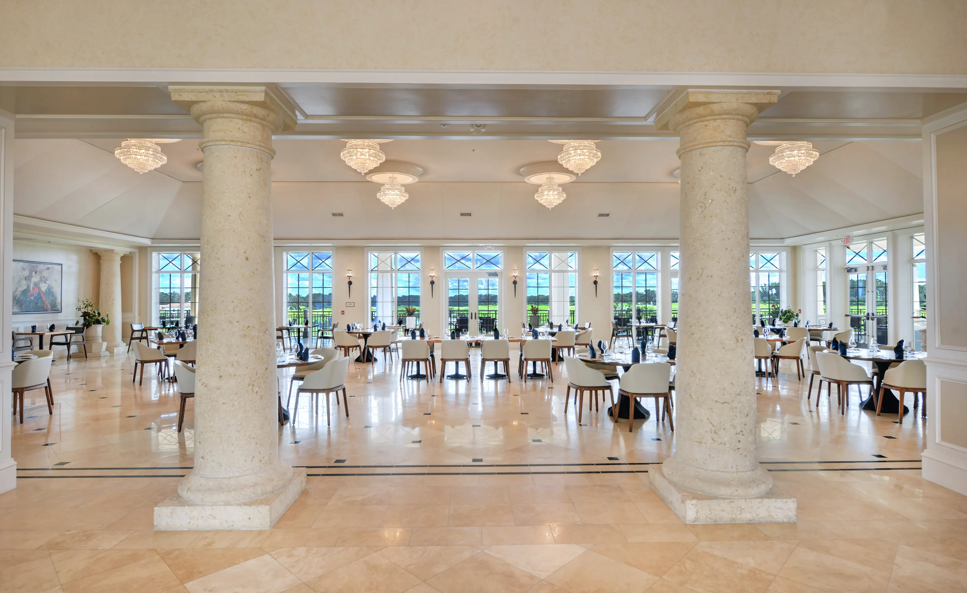 Large dining room in IronCrest with marble tile flooring, stone columns, multiple crystal chandeliers, and wall-to-wall windows.