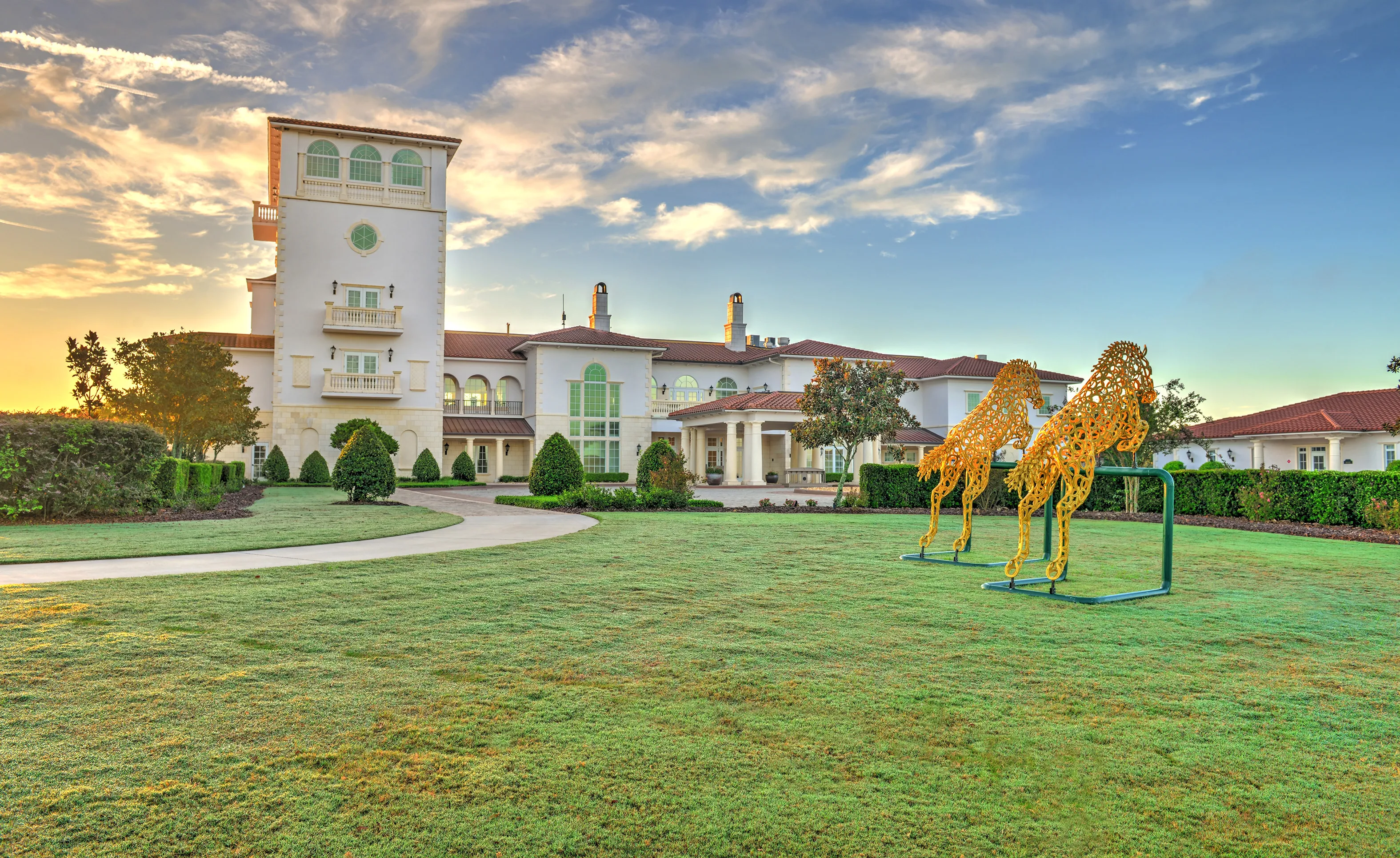 Large multi-story home in IronCrest with white stucco, tile roof, a tall tower, and gold metal horse sculptures on the lawn.
