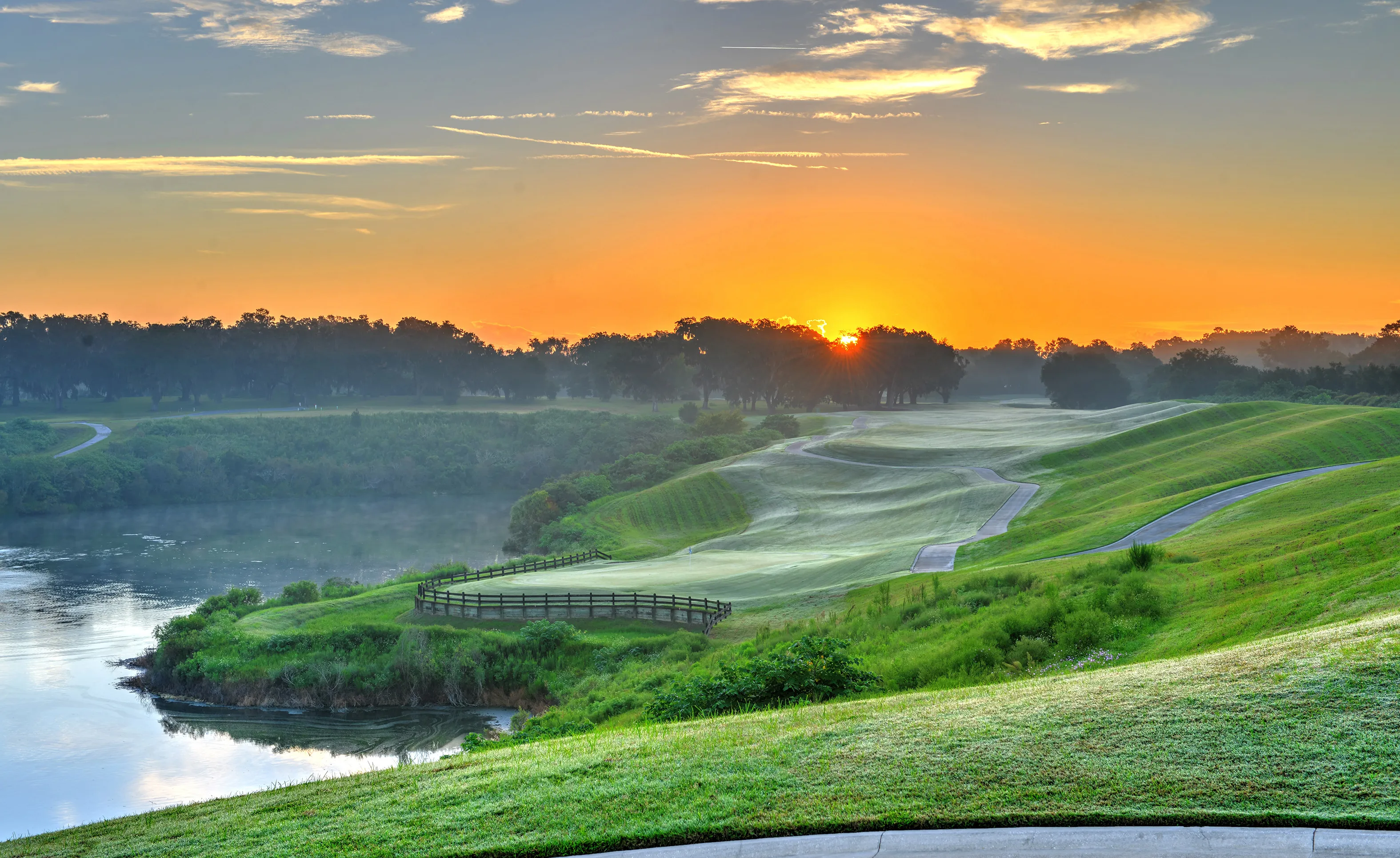 Golf course landscape at IronCrest with a winding concrete path, a pond, and rolling green hills at sunrise.