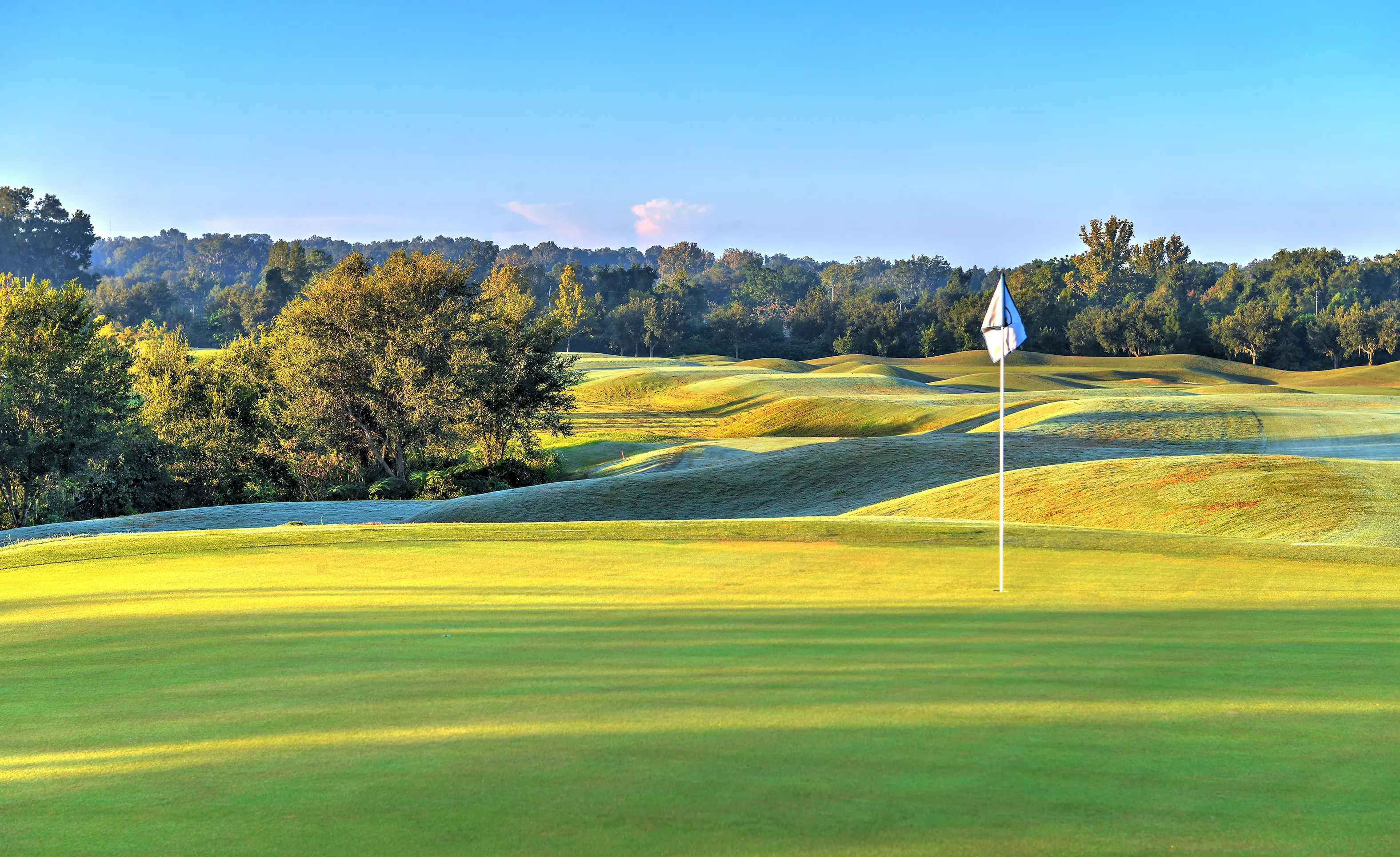 Green golf course fairway at IronCrest with rolling mounds, a flagstick, and a backdrop of dense trees under a clear sky.