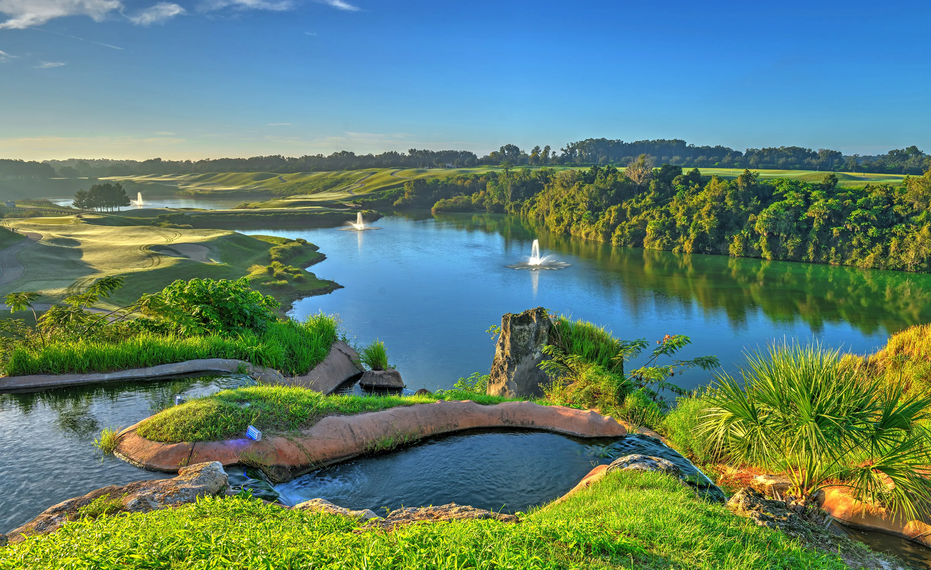 Scenic view of a lake and golf course at IronCrest with cascading water features and lush landscaping under a blue sky.