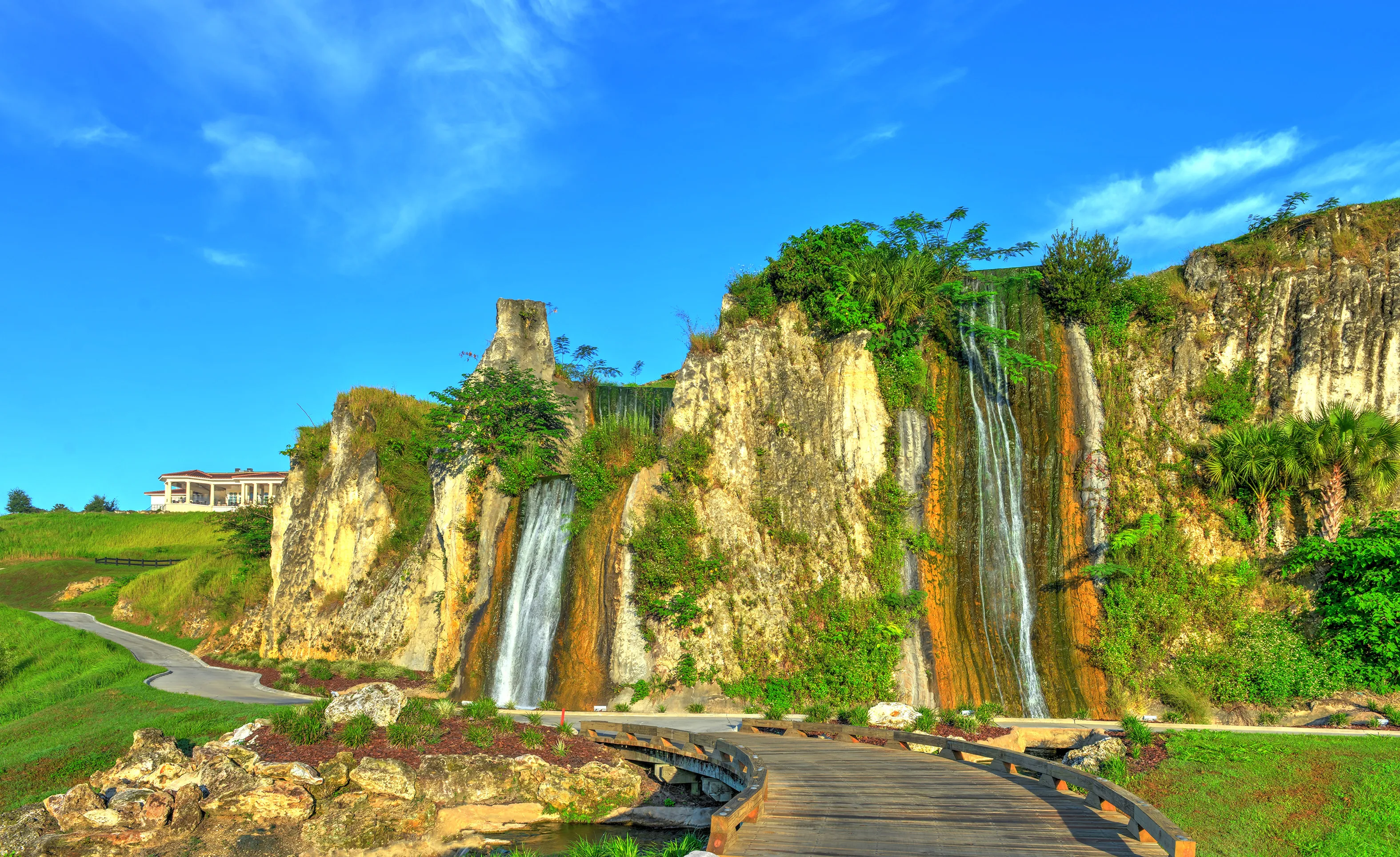 Waterfalls cascade down a natural stone cliff with a wooden footbridge in the foreground at the IronCrest community.