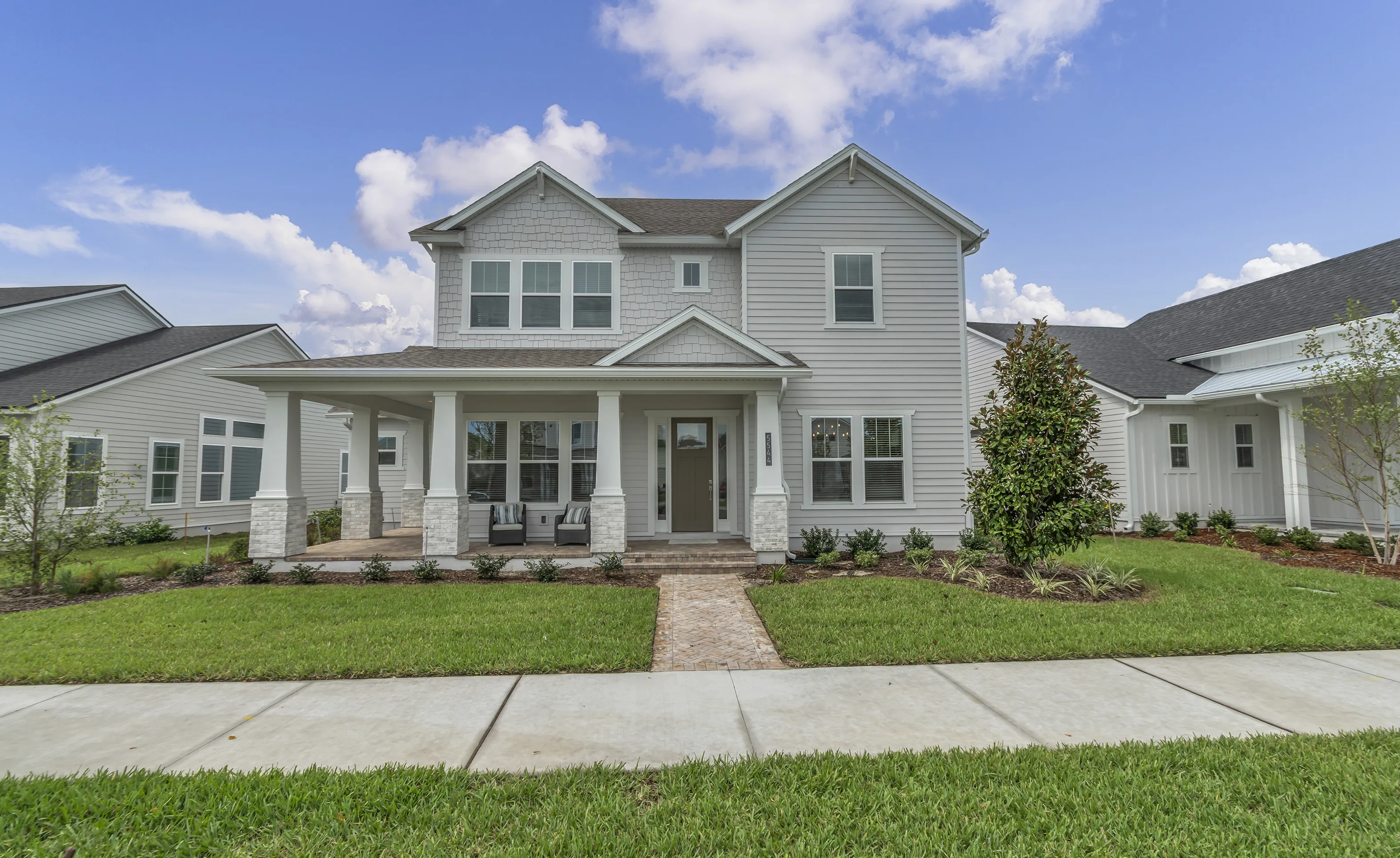 Front exterior of a two-story gray home with a covered front porch, stone-based columns, and a brick paver walkway.