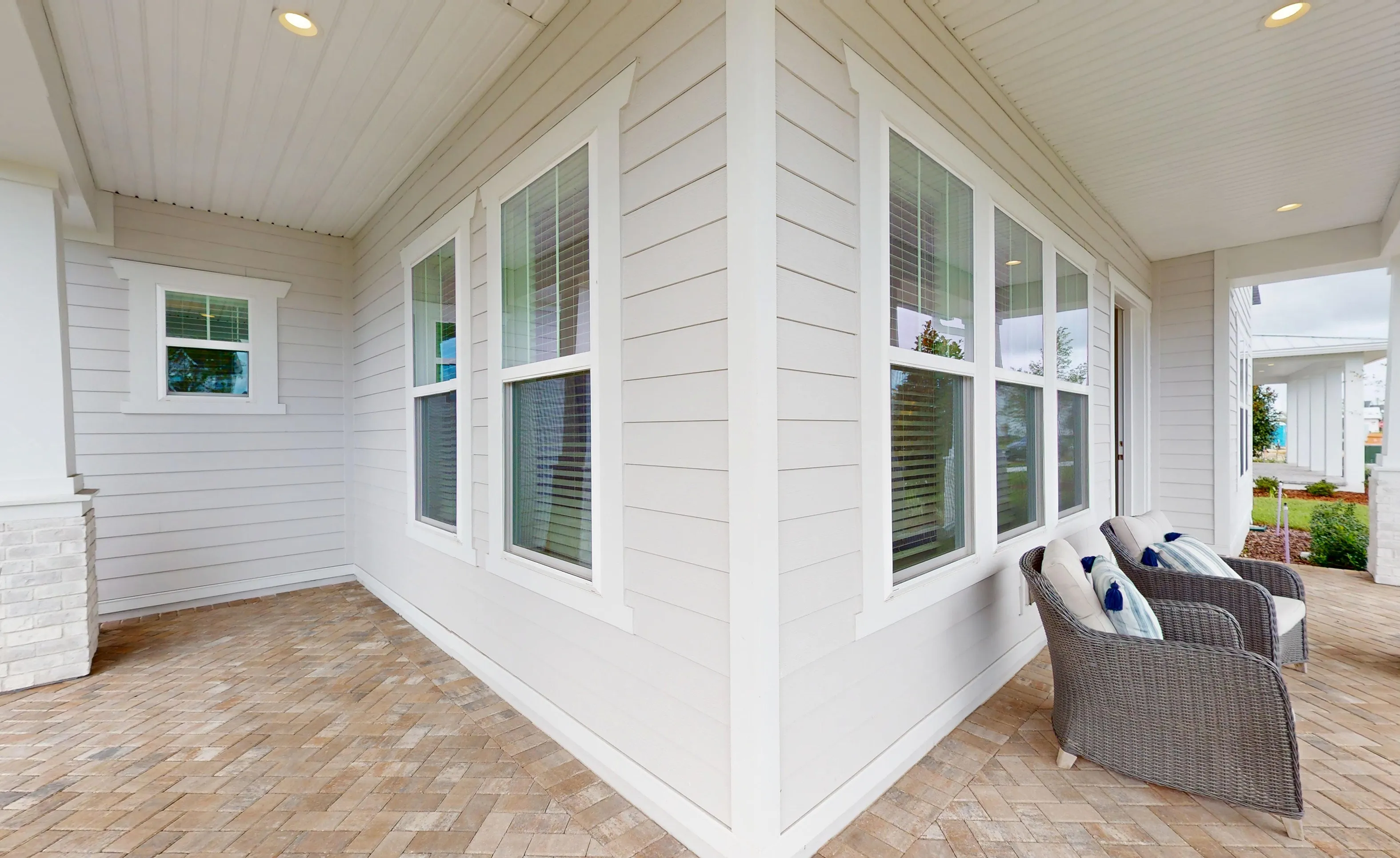 Covered porch at Braddock plan with light horizontal siding, paver flooring, white trim, and gray wicker patio chairs.