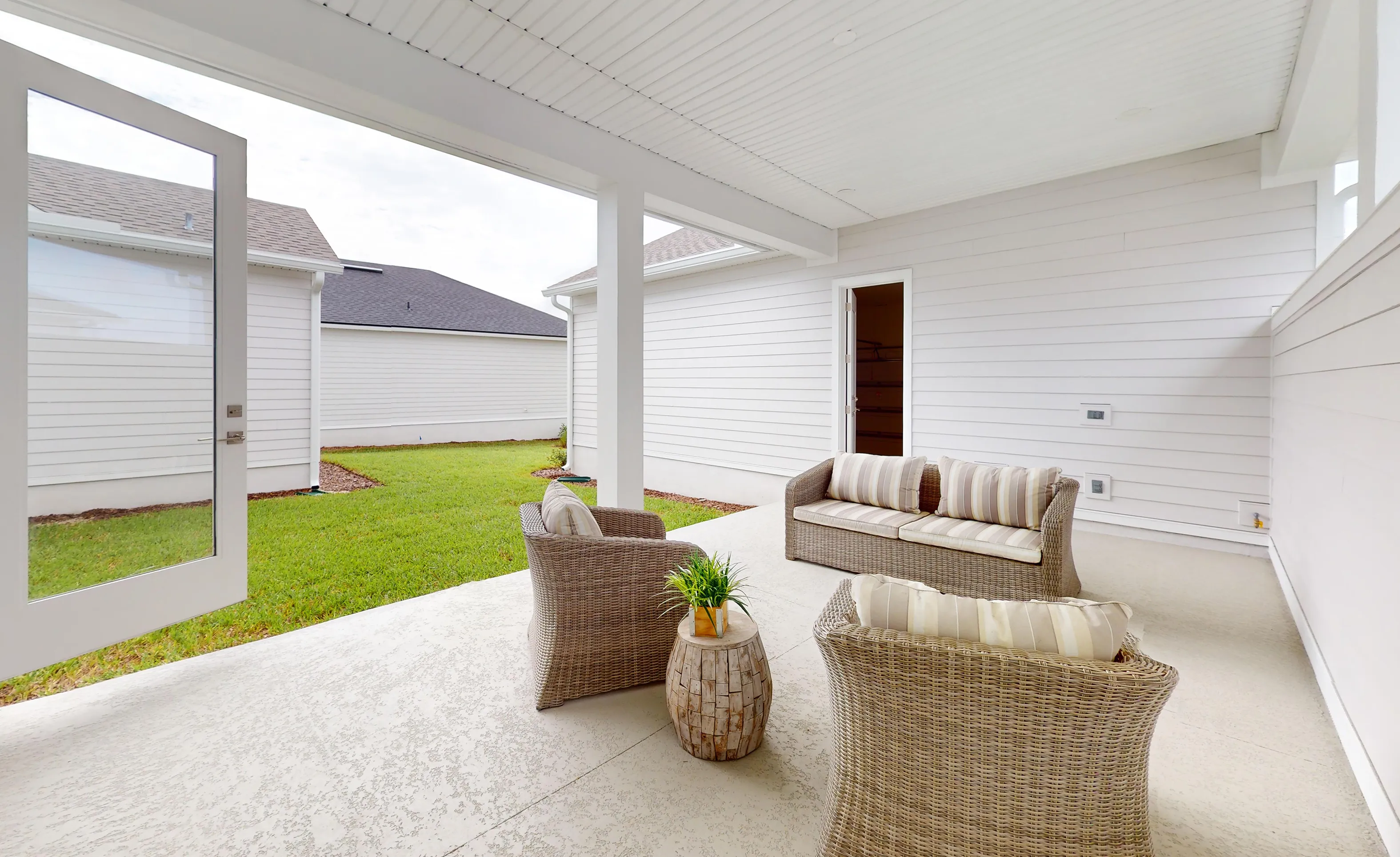 Covered porch of the Braddock plan with white siding, concrete floor, wicker furniture, and an open door to the backyard.