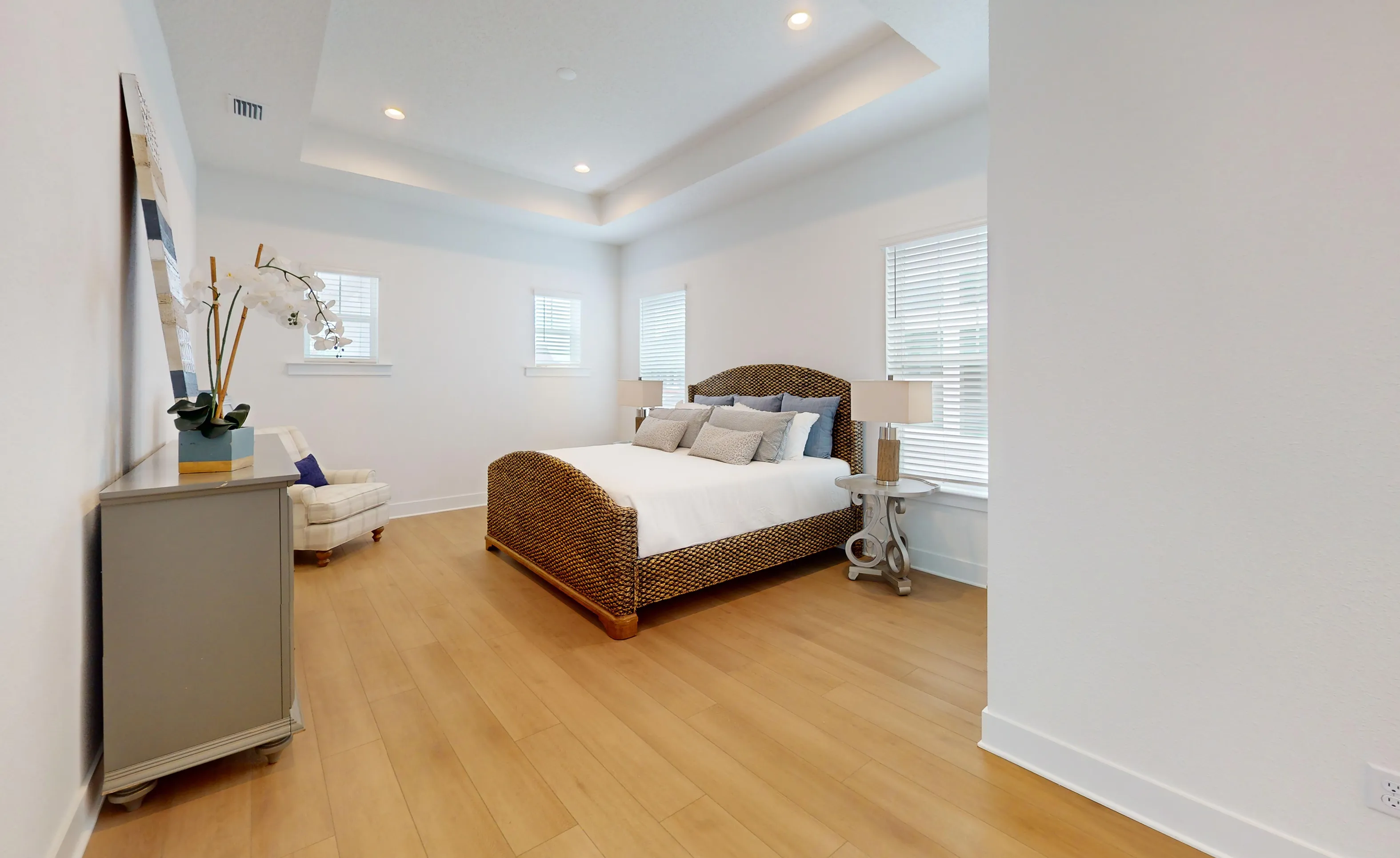 Primary bedroom in the Braddock plan with wood flooring, tray ceiling, woven bed frame, and large windows with blinds.