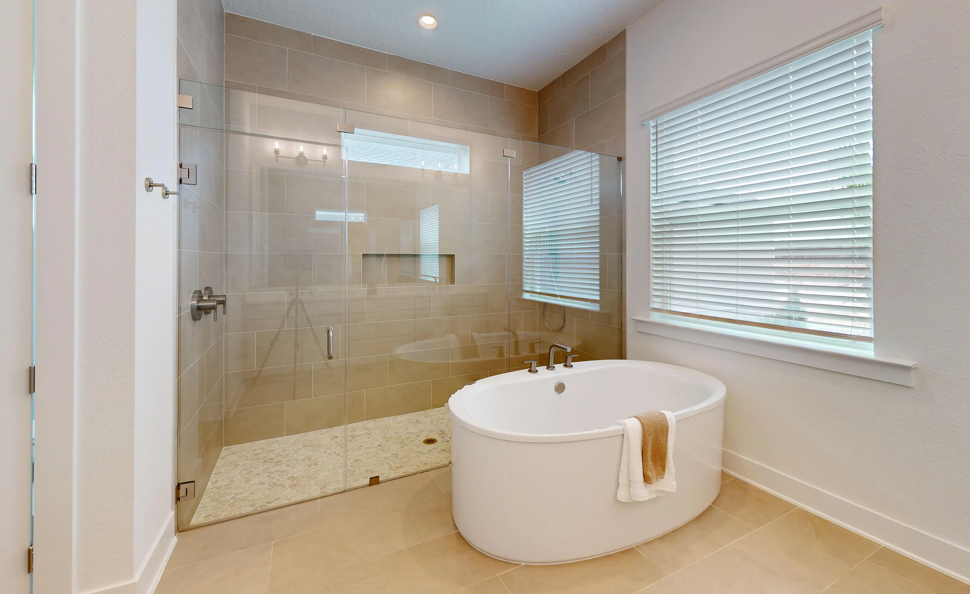 Primary bath with a white oval soaking tub, large glass-enclosed shower with tiled walls, and a window with blinds.