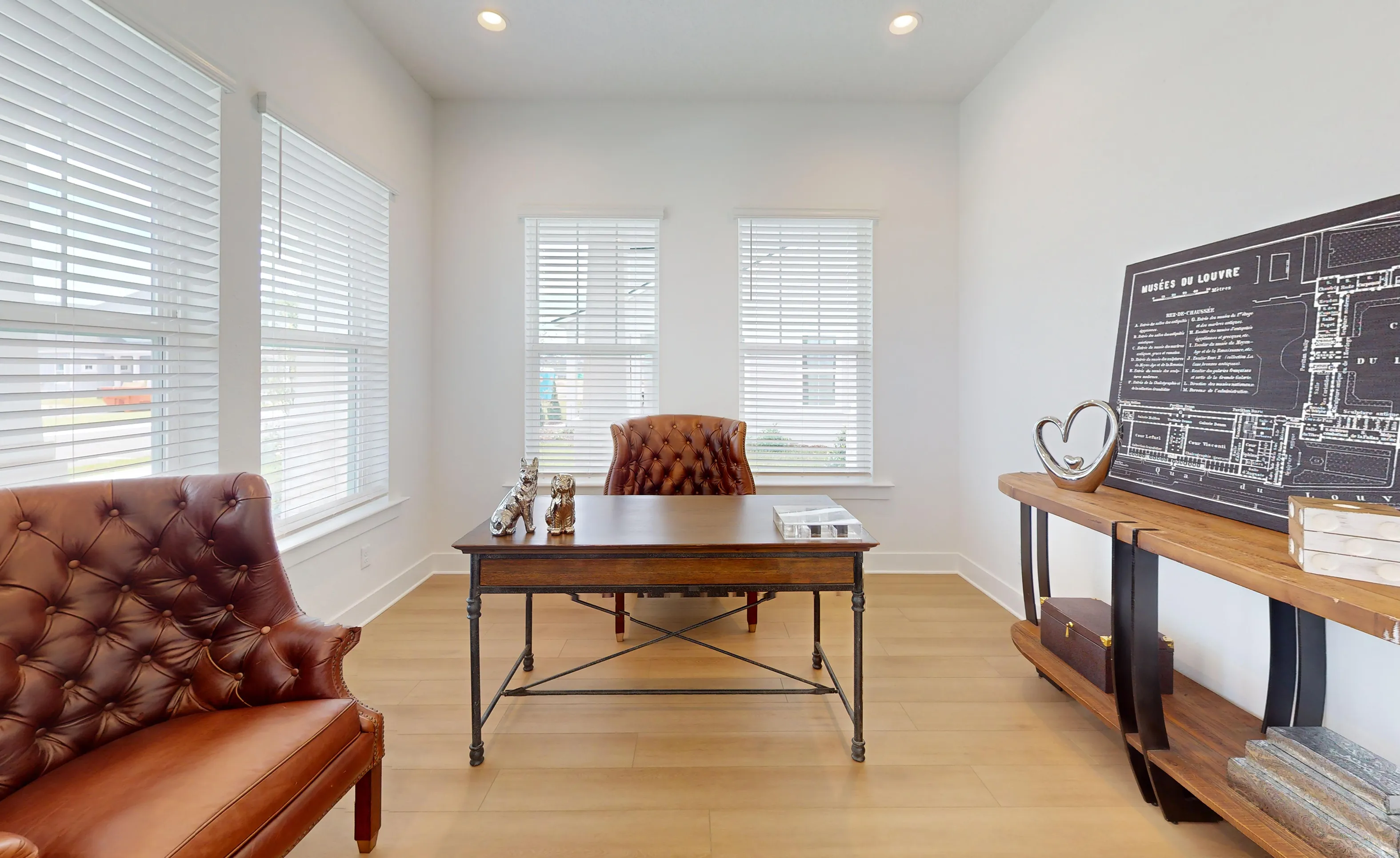 Home office in the Braddock plan with wood-look floors, white walls, four windows with blinds, a wood desk, and leather chairs.