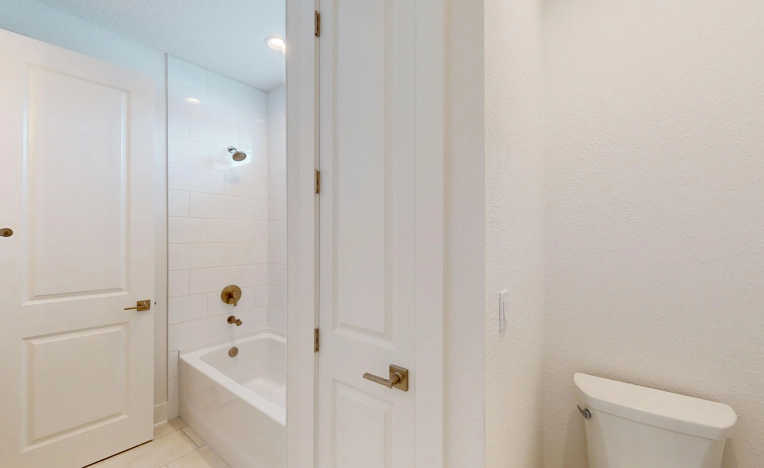 Secondary bath with white subway tiled tub-shower combo, gold-toned fixtures, and a toilet in a white-walled room.